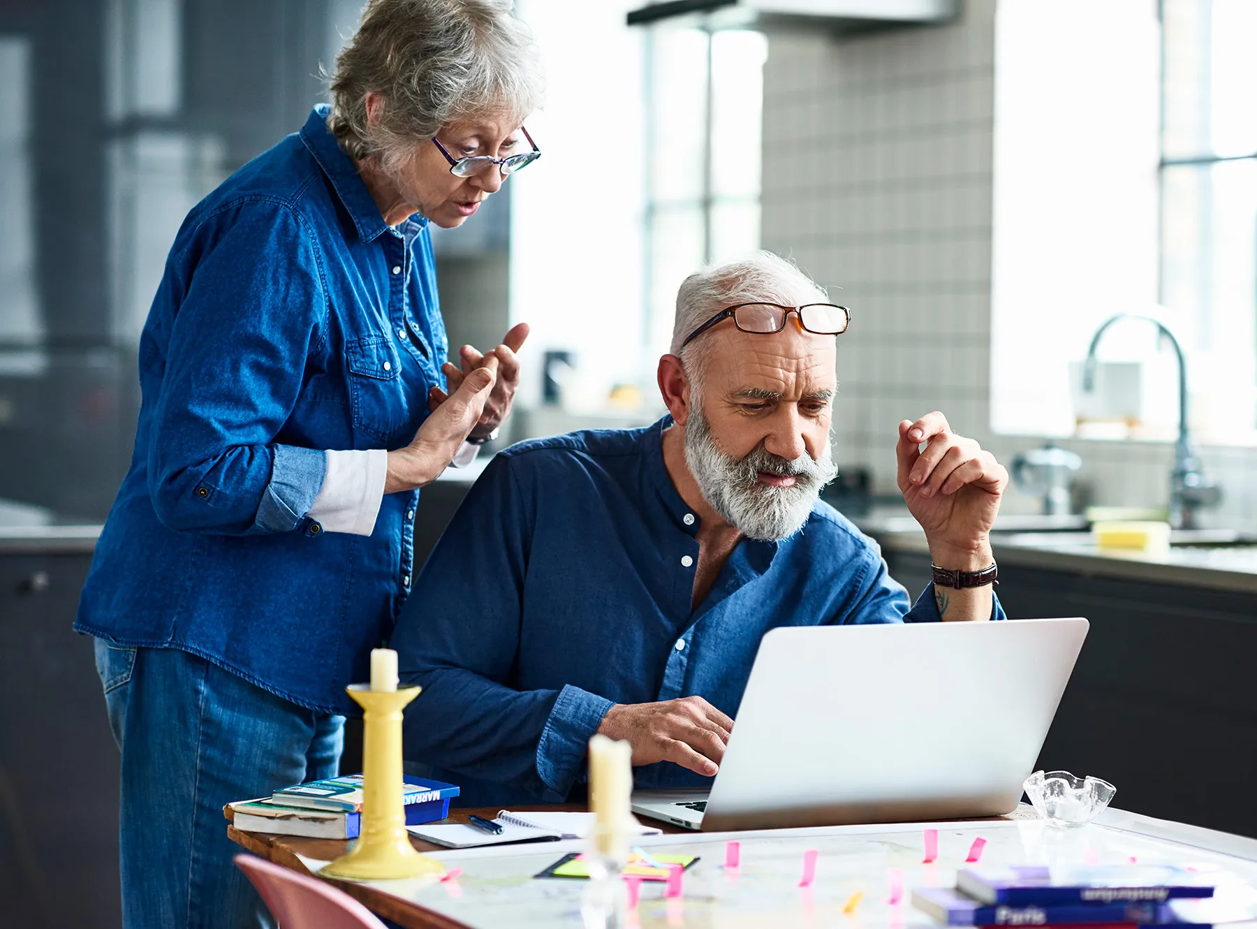 Older couple planning travel at a kitchen table, with the man using a laptop and the woman looking over his shoulder.