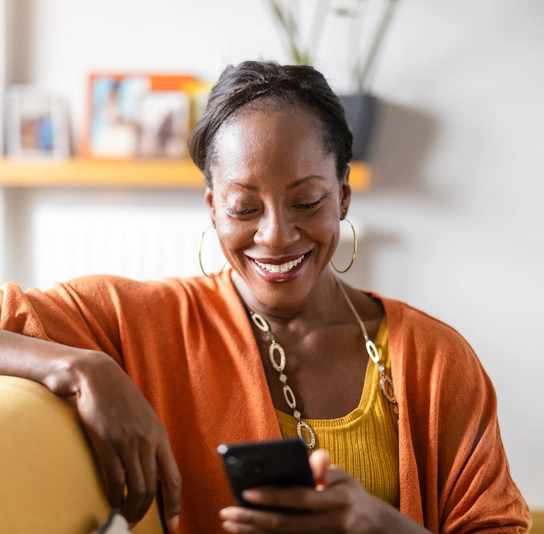 Smiling woman in a cozy home environment looking at her smartphone while sitting on a yellow couch.