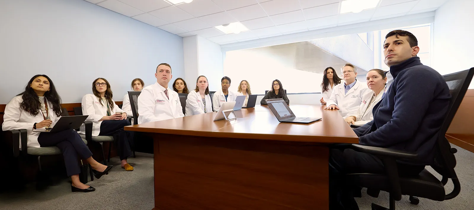 A team of physicians in white coats and healthcare staff sit around a large conference table, engaged in a tumor board meeting with laptops open.