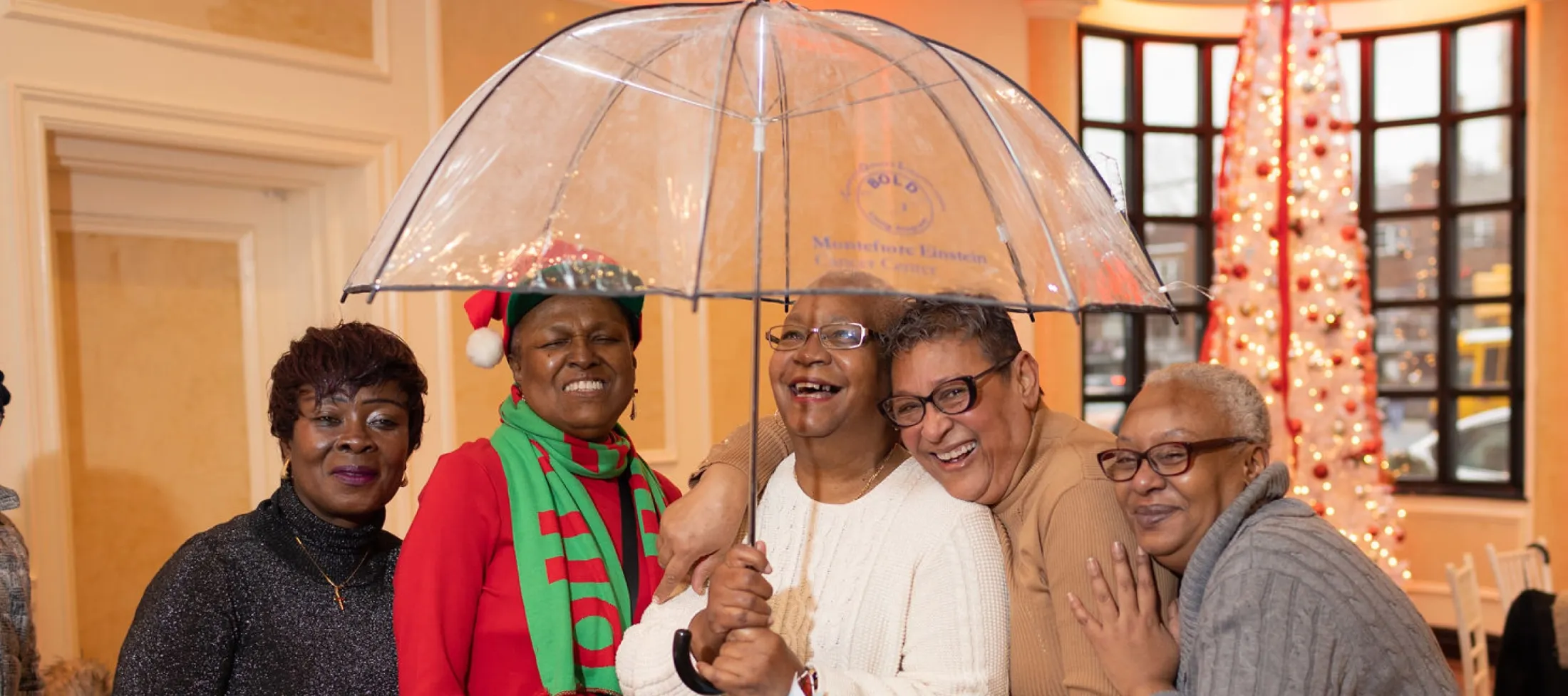 Five BOLD party attendees laughing together under a large, clear umbrella indoors