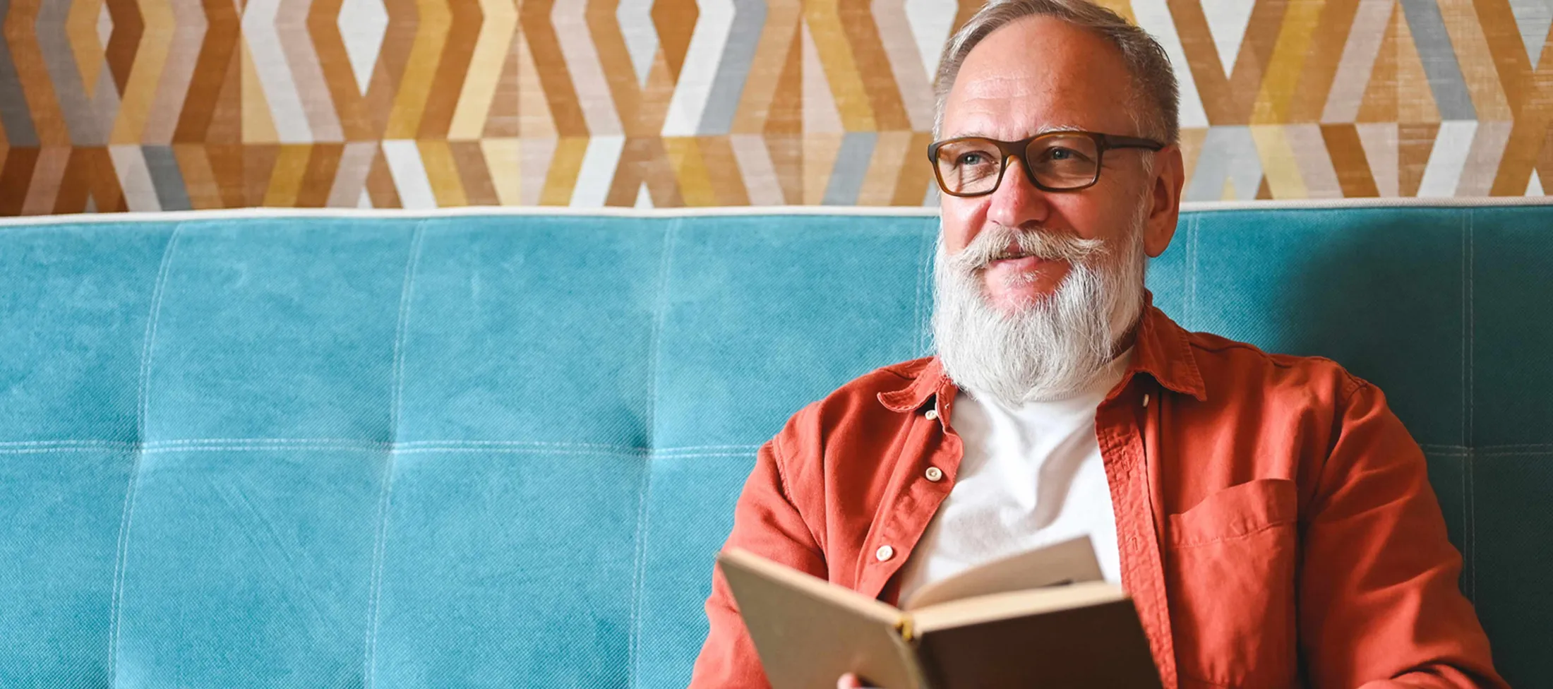 Man with beard and wearing glasses reading a book while seating on a blue couch