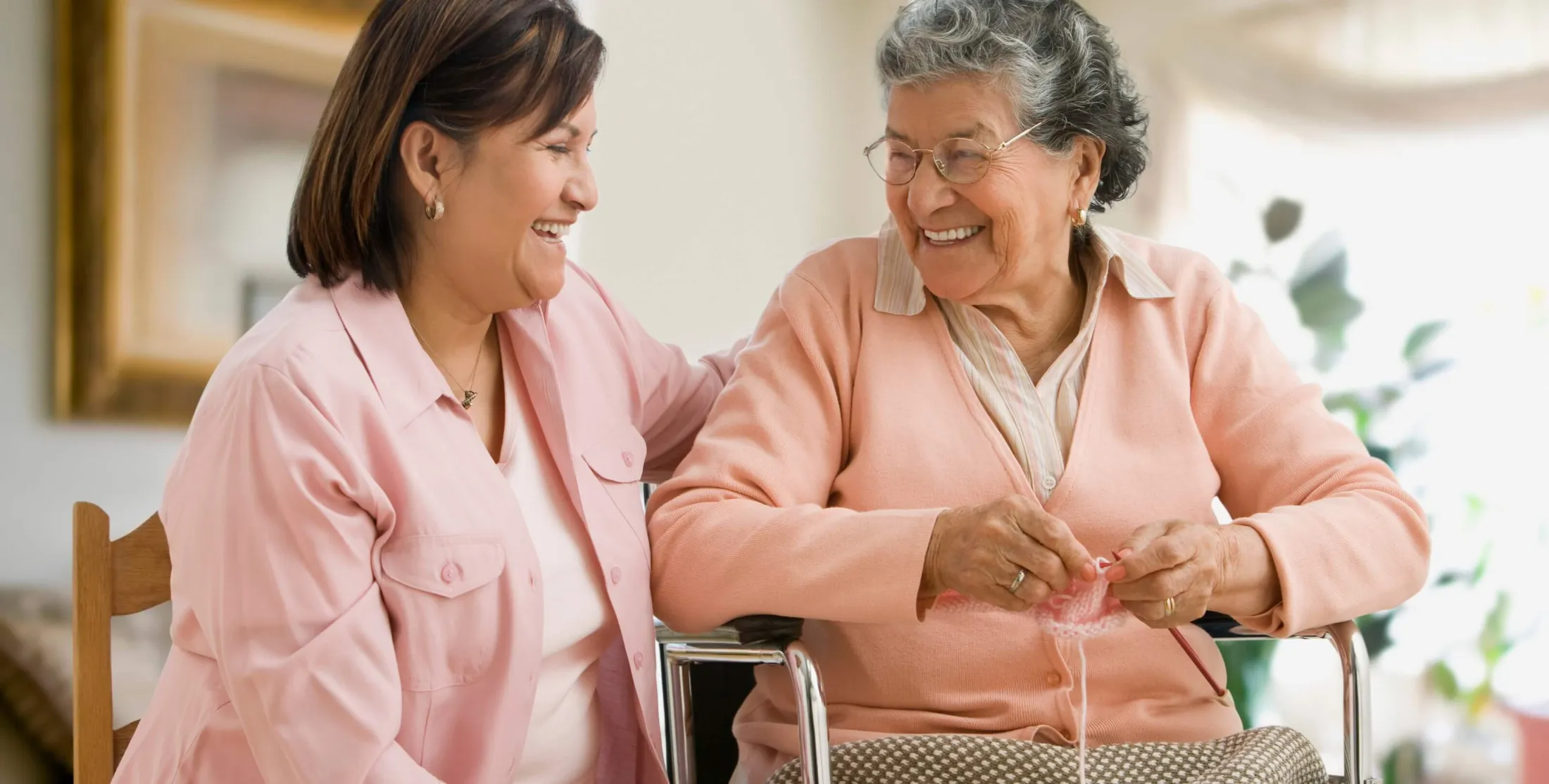 A woman sits beside an older woman in a wheelchair, both appearing happy and engaged in conversation