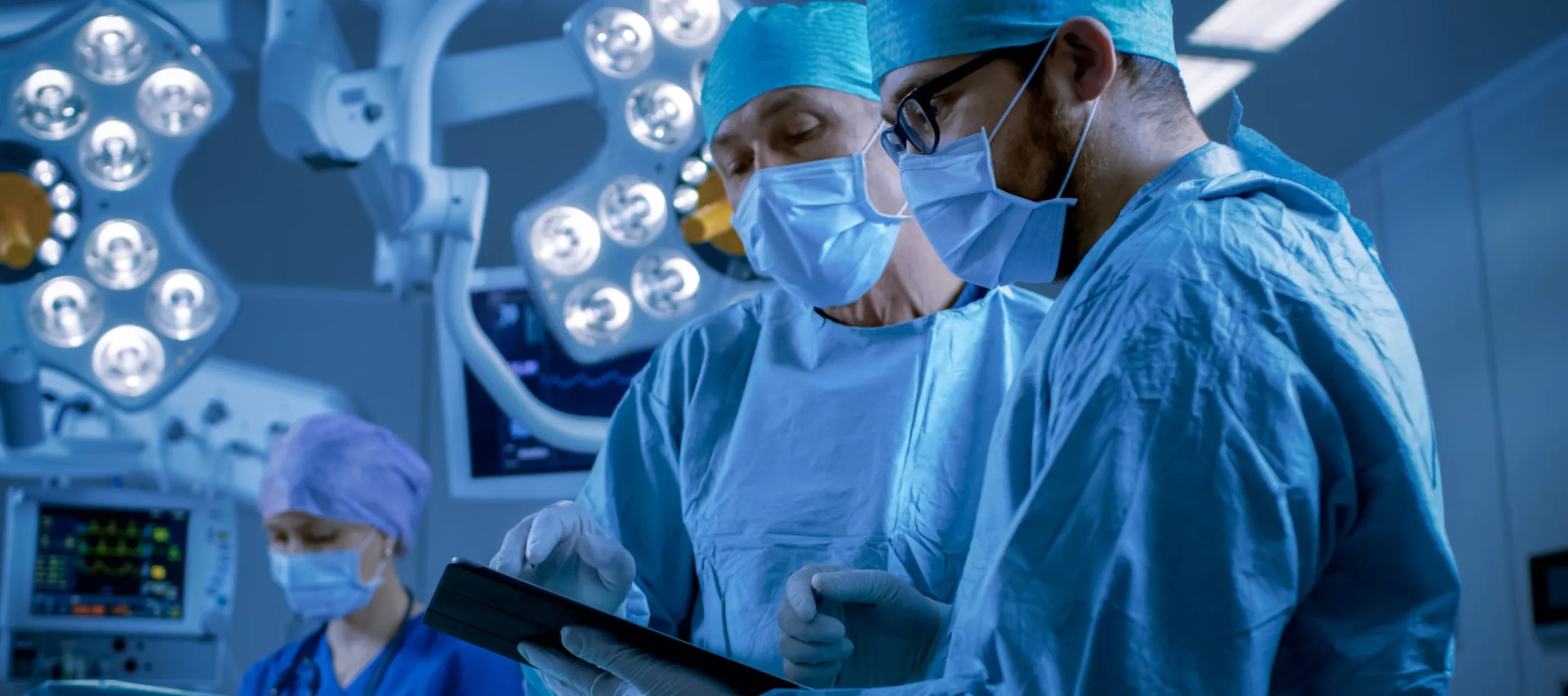 Two surgeons wearing blue surgical gowns, caps, face masks, and sterile gloves stand together in a blue-lit operating room while reviewing data on a tablet device. Bright overhead surgical lights and a patient monitor screen are visible in the background.