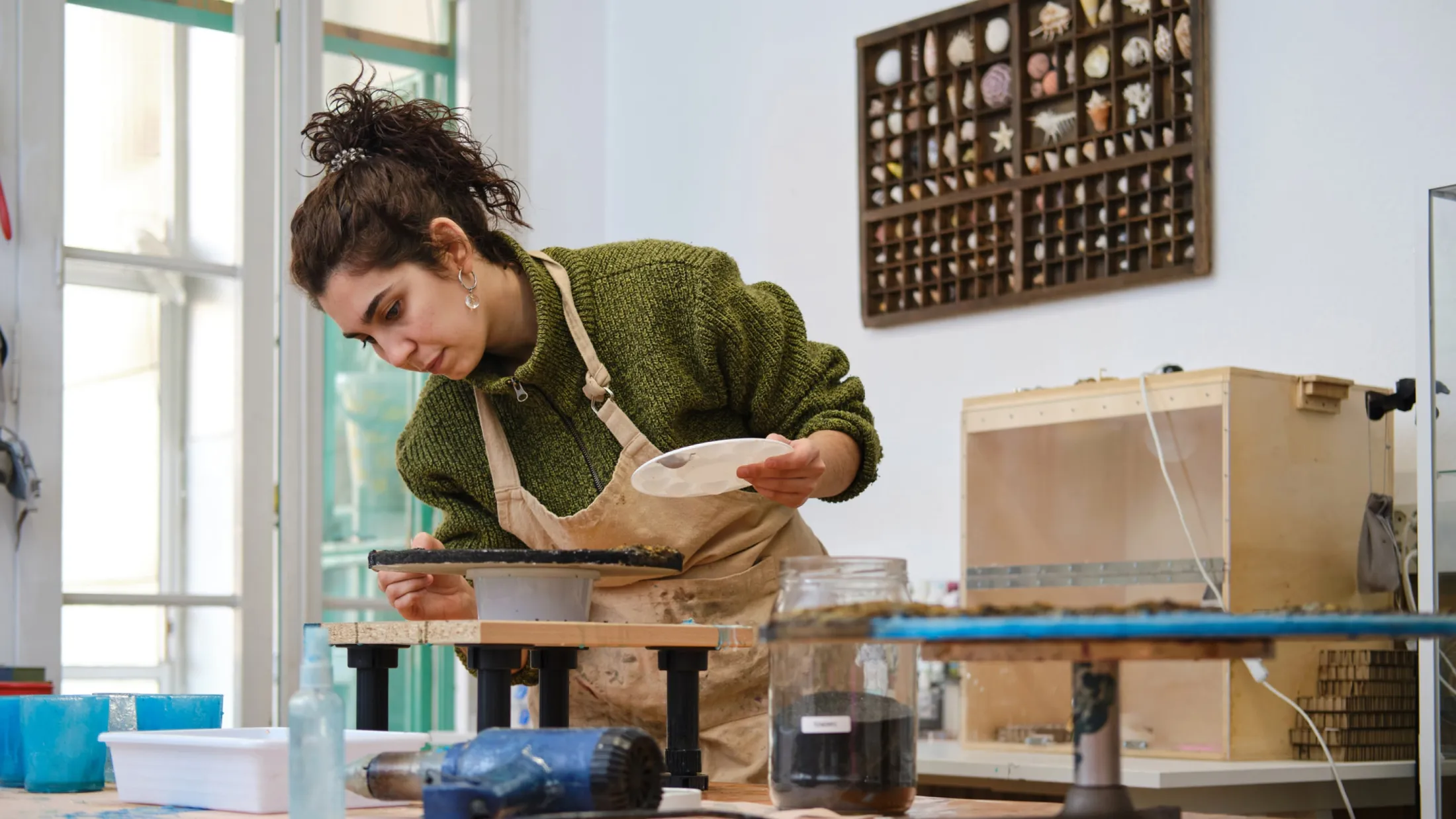 An artist with dark curly hair tied up in a high bun wears a green knit sweater and a beige apron while working in her studio. She leans closely over a piece of pottery set on a small turntable, holding a paint palette in one hand and carefully applying detail with a brush in the other. In the background, a wall display filled with seashells and various crafting supplies are visible on the workbench.