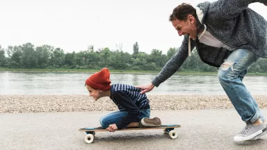 A happy father pushing son on skateboard by a river.