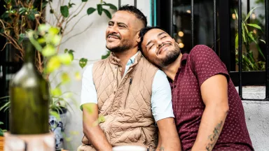 Two bearded men sit together on a bench in a garden enjoying a sunny day. One man's head is resting on the other's shoulder.
