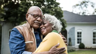 Senior couple smiling while embracing outdoors in front yard of home.