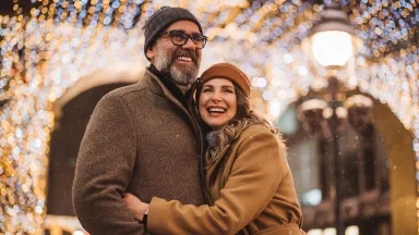 Smiling couple in winter coats hugging under festive holiday lights on a snowy evening