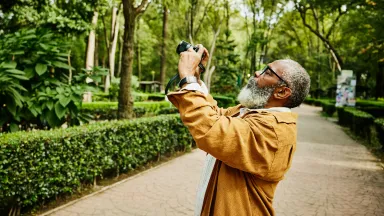Elderly man with a gray beard and glasses taking a photo with a camera in a lush, green park.