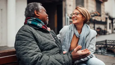 Smiling man and woman sitting on a bench outdoors, warmly dressed and engaged in a lively conversation.