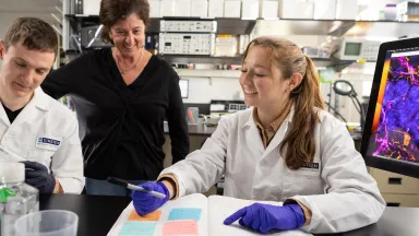 Two students in white lab coats working with lab equipment while a professor observes and smiles.