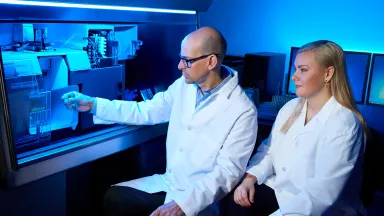 Male and female researchers in lab coats examining samples inside high-tech lab equipment under blue lighting.