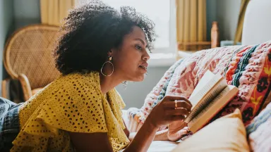 Woman in a yellow top lying on a bed and reading a book in a cozy, sunlit room with colorful bedding.