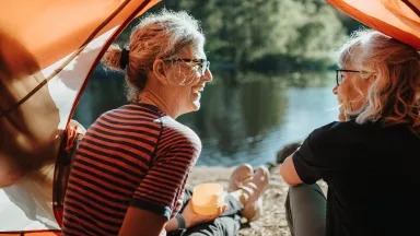  Two older women smiling and talking while sitting at the edge of a tent by a lake, enjoying a peaceful moment in nature.