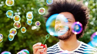 A man smiling while blowing bubbles outside.
