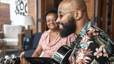 Man playing guitar and smiling while a woman sits beside him, listening happily.