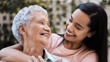 Smiling older woman being embraced by a younger woman, both sharing a joyful moment outdoors.