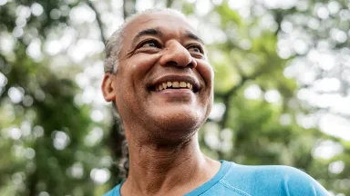 Close-up of a smiling man in a blue shirt outdoors, looking up with sunlight filtering through the trees behind him.