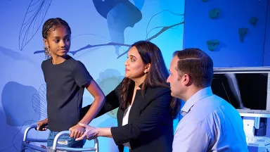 Smiling older woman walking arm-in-arm with a younger woman outdoors beneath leafy trees.