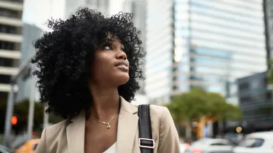 Confident woman with curly hair looking upward while walking through a city street, with modern glass skyscrapers in the background.