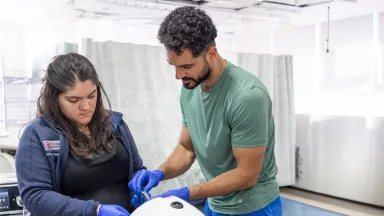 Two people in a clinical simulation center work together on a medical training device, wearing gloves and focused on a hands-on procedure.