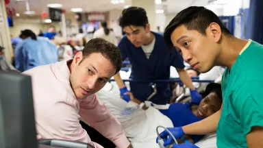 Physicians and residents crowd around a patient in a busy emergency department, intently focused on an ultrasound screen.