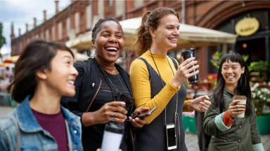 A group of four young women walk together outdoors, smiling and laughing while holding coffee cups, enjoying a lively conversation in an urban setting.