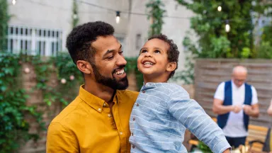A father smiling as he holds his young son outdoors during a family gathering in a backyard
