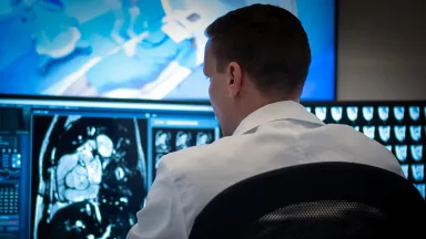 A medical professional wearing a crisp white coat sits in an office chair, viewed from behind as they analyze glowing diagnostic scans. They face multiple computer monitors displaying detailed MRI, CT scans, and medical imaging data, with a larger display screen visible in the background above.