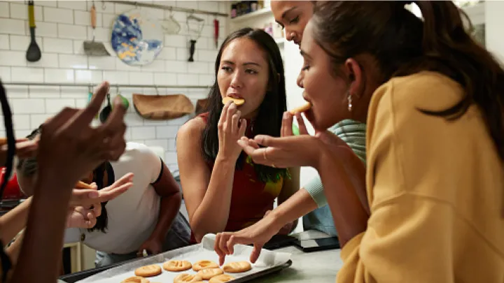 A group of young women of various backgrounds gather around a tray of fresh cookies in a kitchen with white tile walls.