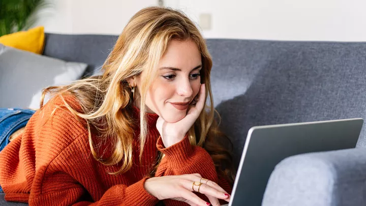 Smiling woman in orange sweater lying on a couch, using a laptop in a cozy, modern living room.