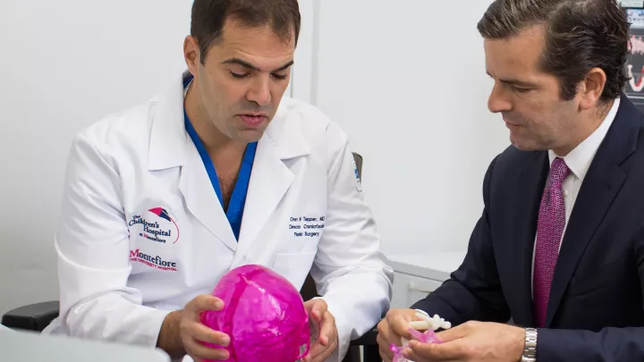 Two men, one in a white lab coat and one in a suit, examine 3D-printed skull models in a medical office setting.