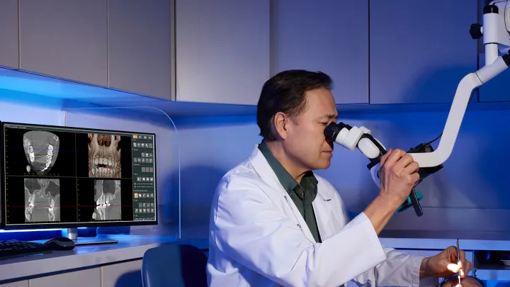 Dentist using a surgical microscope while examining a patient, with dental X-rays displayed on a monitor.