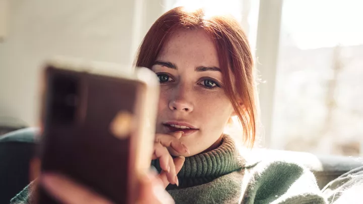 Woman with red hair in a cozy sweater looking at her phone indoors.