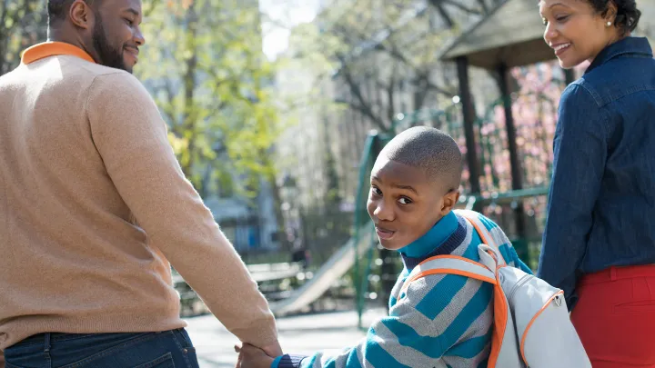 Young boy with backpack holds hands with parents while heading to school in a sunny park.