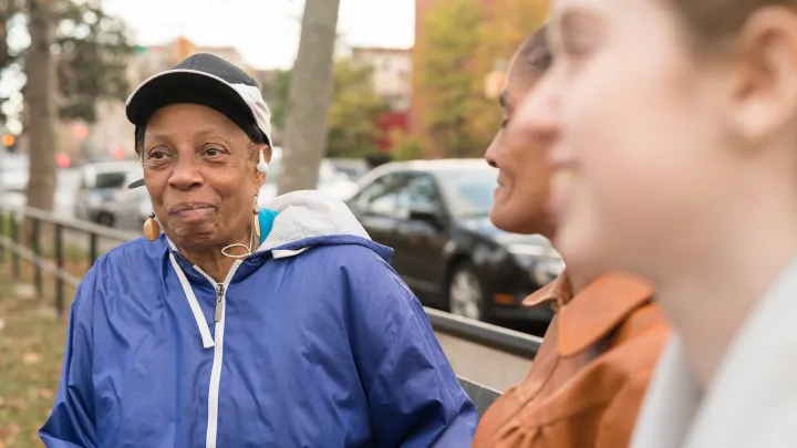 Elderly woman in blue jacket sits on a bench talking with two women in an urban park.