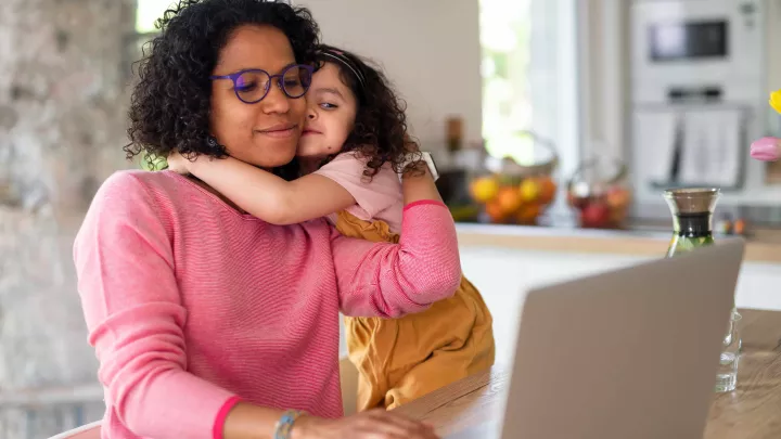 Child embracing her mother while mother is typing on laptop