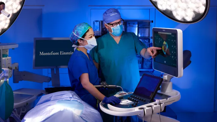 Two medical professionals in surgical attire examining an ultrasound monitor in an operating room, with bright surgical lights and Montefiore Einstein screen in the background.