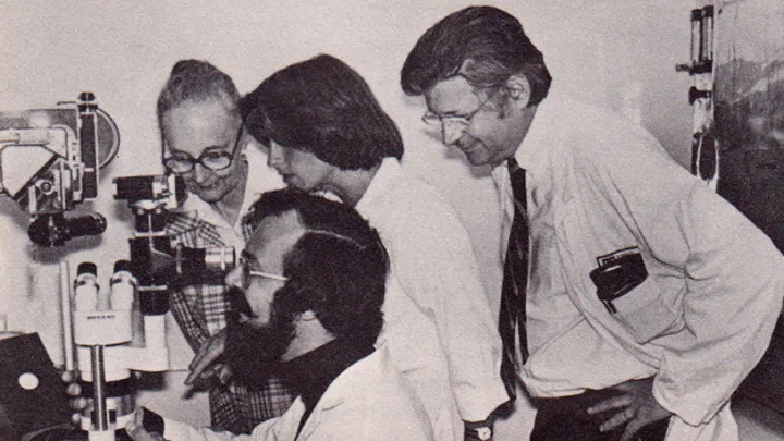 Vintage photo of four medical professionals gathered around a microscope, observing closely and smiling, taken in 1979.