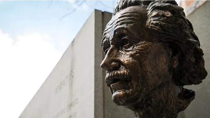 Bronze bust of Albert Einstein mounted outdoors against a concrete wall.