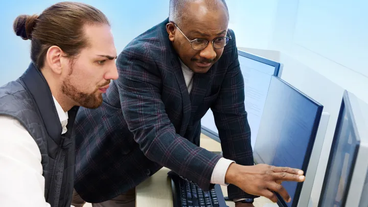 Two men collaborate at a computer workstation, with one pointing at the screen while explaining something to the other.