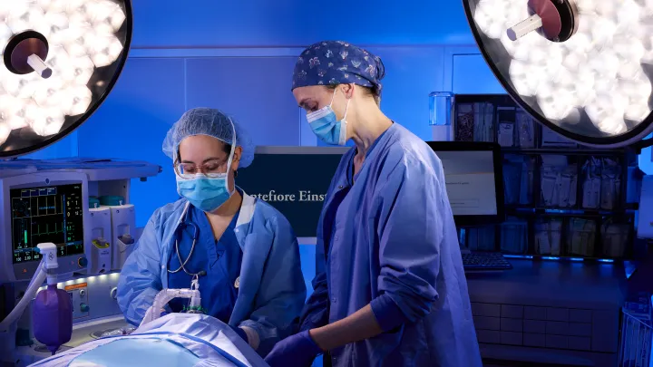 Two medical professionals in surgical scrubs and masks working under bright operating room lights, with a patient on the table and medical equipment in the background.