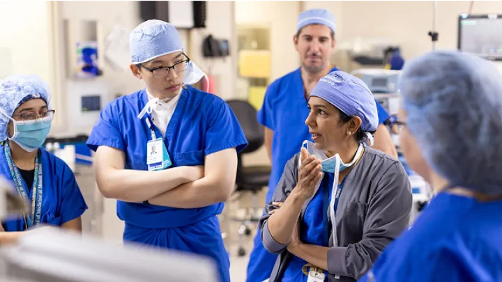 Group of medical trainees and an instructor in discussion during a clinical training session, all wearing scrubs and surgical caps in a hospital setting.