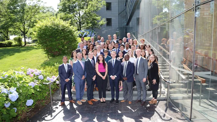 Large group of professionally dressed people posing for a team photo outside a modern building with reflective glass and surrounding greenery.