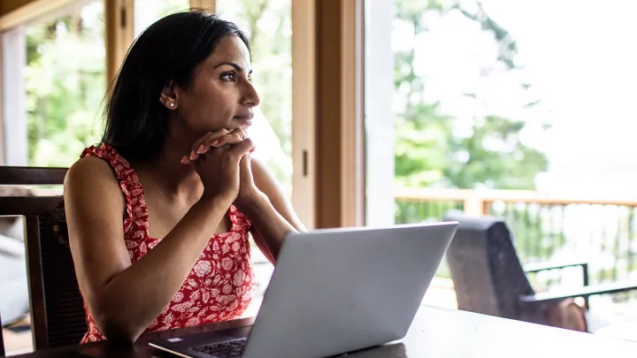 Woman in a red patterned dress sits at a table with a laptop, gazing thoughtfully out a bright window.