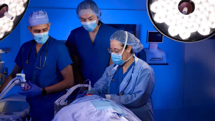 Operating room team in scrubs and masks managing anesthesia for a patient under bright surgical lights.