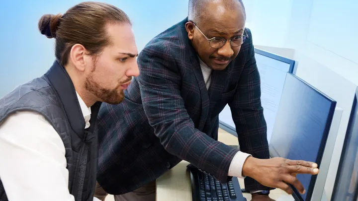 Mentor pointing at a computer monitor while reviewing data with a colleague in an office.