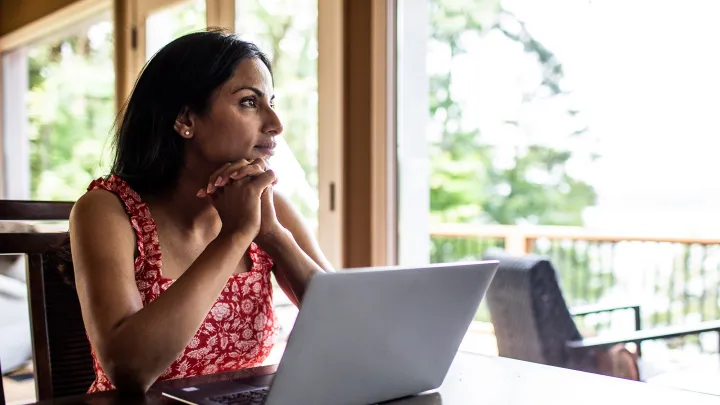 Woman in a red patterned dress sitting at a table with a laptop, looking thoughtfully out a bright window.