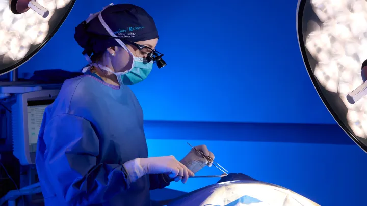 Surgeon in blue scrubs and mask preparing tools under surgical lights.