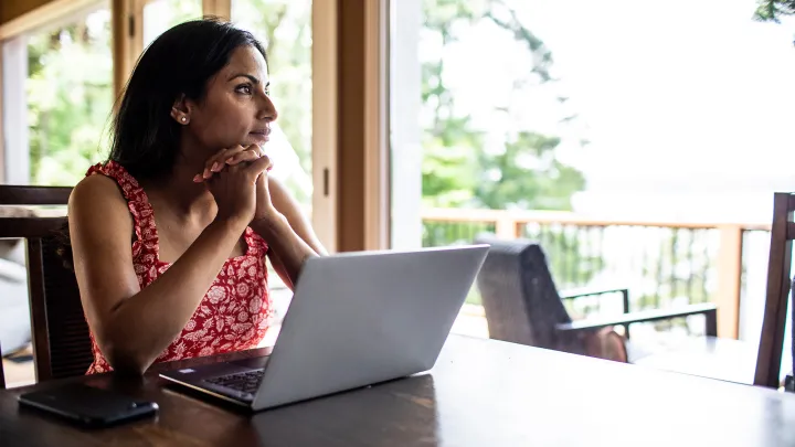 Woman in a red floral dress sitting at a table with a laptop, gazing thoughtfully out a window in a bright, wooded setting.